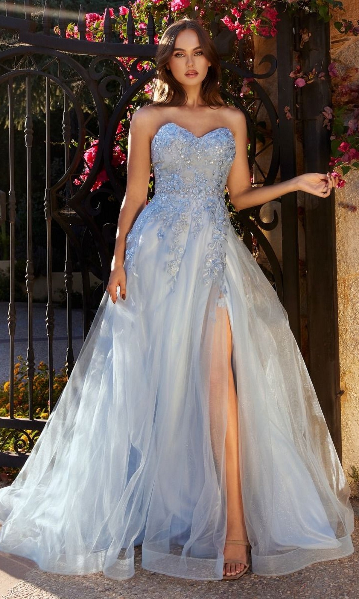 A woman stands before a gate in the Pastel Blue Ball Gown A1339 by Andrea and Leo, featuring a strapless design, floral embellishments, and a high slit, as she holds the side of her dress.